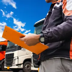 Kierowca z teczką, na której widnieje świadectwo kwalifikacji zawodowej, stoi przed rzędem ciężarówek.
