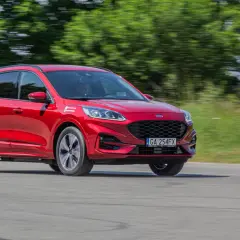 Czerwony Ford Kuga Hybrid w ruchu, z rozmytym tłem zieleni i drogi.