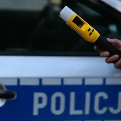 Dłoń trzymająca żółty alkomat na tle radiowozu policyjnego z napisem "POLICJA".