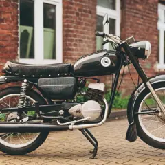 Czarny motocykl WSK 125, z charakterystycznym emblematem na baku, który sugeruje jego pojemność. Stoi na bruku przed ceglanym budynkiem.