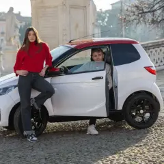 Dwoje młodych ludzi przy białym samochodzie z czerwonym dachem na brukowanej ulicy.