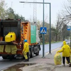 Śmieciarka i dwie osoby w żółtych płaszczach zbierające worki z odpadami.