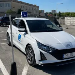 Biały samochód nauki jazdy z literą "L" na dachu, na placu manewrowym.