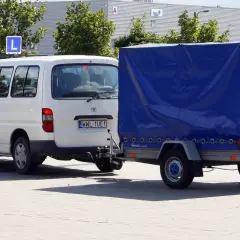 Biały van z przyczepą, na której widnieje literka "L", symbolizuje naukę jazdy z przyczepą, przygotowującą do zdobycia prawa jazdy kategorii B+E.