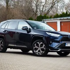 Ciemnoniebieski SUV Toyota RAV4 hybrydowy zaparkowany przed garażami.