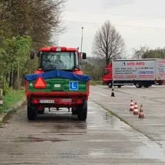 Czerwony traktor z przyczepą na placu manewrowym, obok ciężarówka AUTO-MOTO.