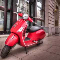 Czerwony skuter Vespa, idealny do zdobycia **prawo jazdy AM**, stoi na chodniku przed budynkiem.