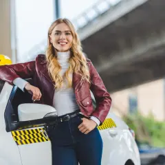 Uśmiechnięta blondynka w skórzanej kurtce opiera się o białe taxi. Chcesz wiedzieć, jak zostać kierowcą Opti Taxi?