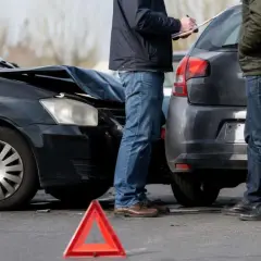 Dwaj mężczyźni spisują protokół po kolizji. Zniszczona maska czarnego auta i czerwony trójkąt ostrzegawczy sugerują, że czeka ich mandat za kolizje.