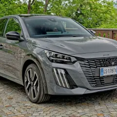 Szary Peugeot 208 zaparkowany na brukowanej drodze, z zielenią w tle.
