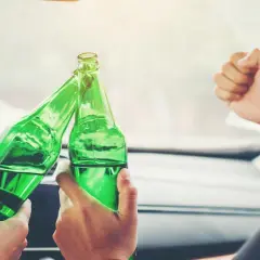 Dwie osoby w samochodzie wznoszą toast zielonymi butelkami piwa. Zastanawiają się, ile można wypić alkomat.