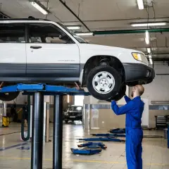 Mechanik w niebieskim kombinezonie naprawia podniesiony samochód, być może usuwając przyczynę piszczących hamulców.