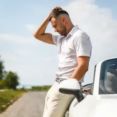 Zamyślony mężczyzna opiera się o biały kabriolet na poboczu drogi. Zastanawia się, kto traci zniżki kierowca czy właściciel.