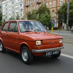 Pomarańczowy Fiat 126p jedzie ulicą. Jego pojemność baku to 25 litrów, co pozwala na długie podróże.