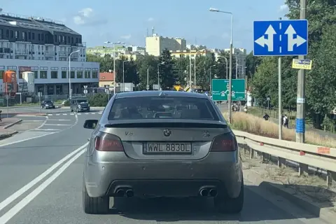 Szare BMW z rejestracją WWL 8830L na drodze, w tle budynki i znaki drogowe.