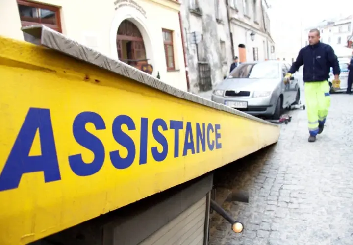 Żółta platforma lawety z napisem "ASSISTANCE" w niebieskich literach. W tle mężczyzna w jaskrawej kamizelce pomaga przy samochodzie.