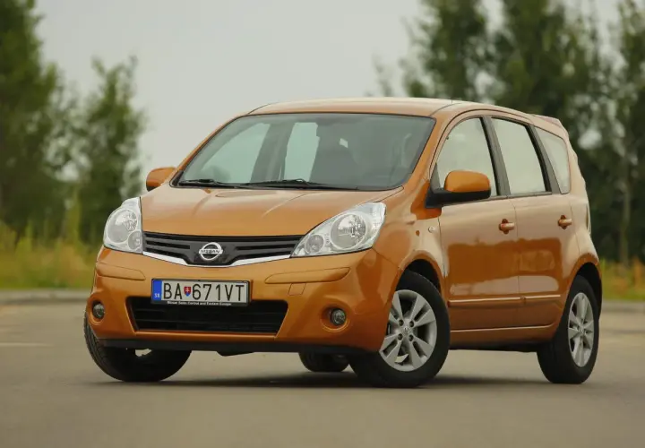 Pomarańczowy Nissan Note z przodu, na tle drzew.