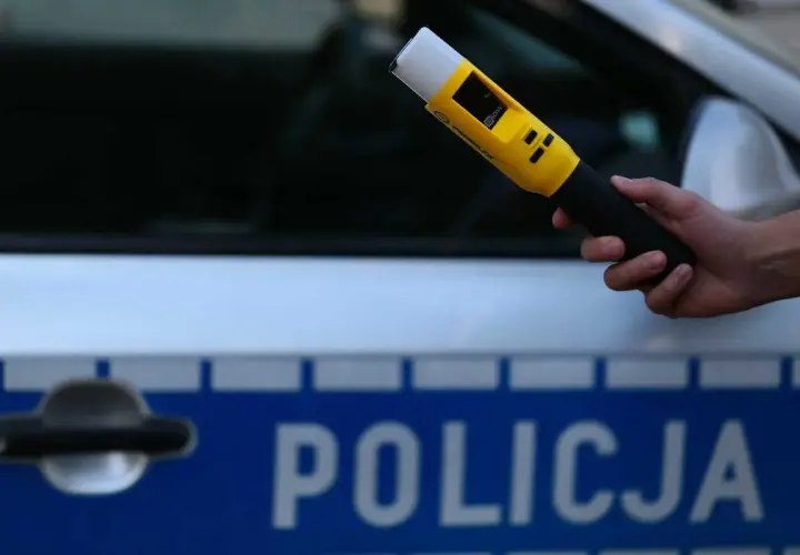 Dłoń trzymająca żółty alkomat na tle radiowozu policyjnego z napisem "POLICJA".