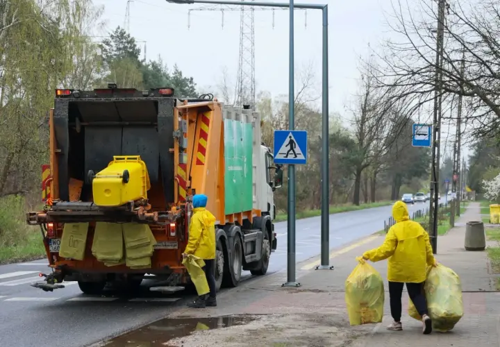 Śmieciarka i dwie osoby w żółtych płaszczach zbierające worki z odpadami.