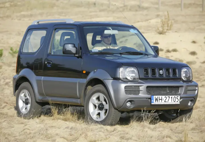 Czarny Suzuki Jimny na piasku. Kierowca w okularach. Pojemność baku tego modelu jest idealna na długie wyprawy.