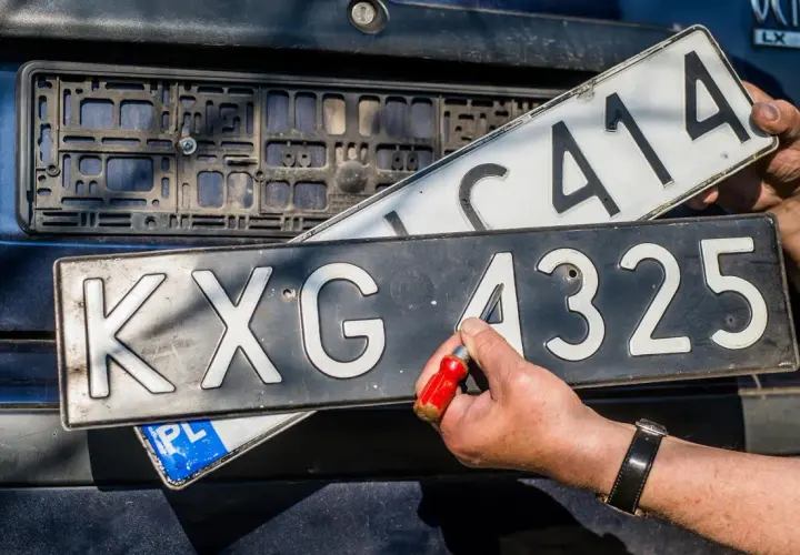 Wymiana tablic rejestracyjnych. Jedna tablica z numerem KXG 4325 jest przykręcana, druga z numerem C414 jest trzymana w ręku.