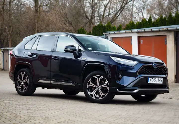Ciemnoniebieski SUV Toyota RAV4 hybrydowy zaparkowany przed garażami.