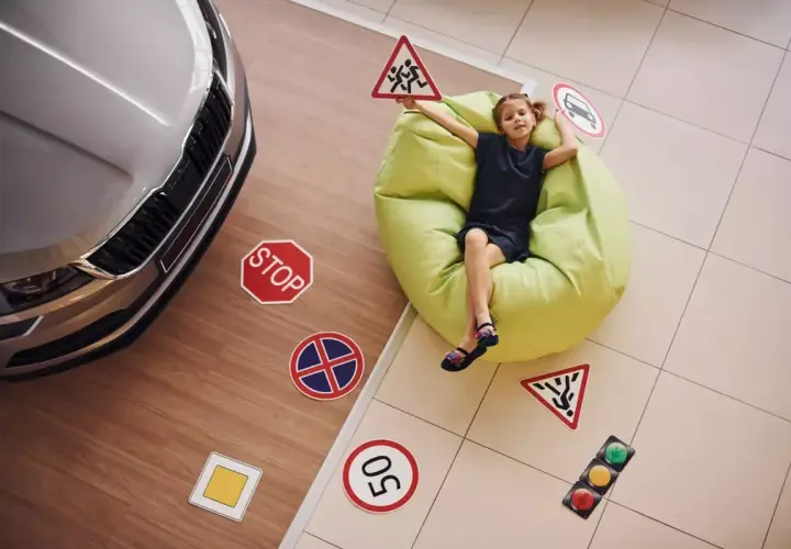 Dziewczynka z tabliczkami drogowymi, jakby uczyła się, ile jest godzin jazdy na prawo jazdy. Obok samochód i znaki: stop, dzieci, ograniczenie prędkości.