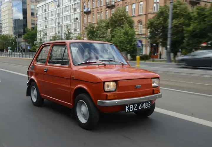 Pomarańczowy Fiat 126p jedzie ulicą. Jego pojemność baku to 25 litrów, co pozwala na długie podróże.