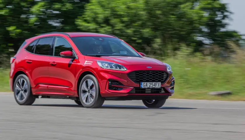 Czerwony Ford Kuga Hybrid w ruchu, z rozmytym tłem zieleni i drogi.