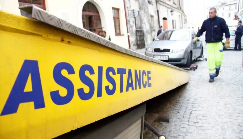 Żółta platforma lawety z napisem "ASSISTANCE" w niebieskich literach. W tle mężczyzna w jaskrawej kamizelce pomaga przy samochodzie.