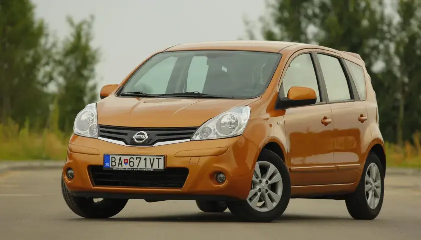 Pomarańczowy Nissan Note z przodu, na tle drzew.