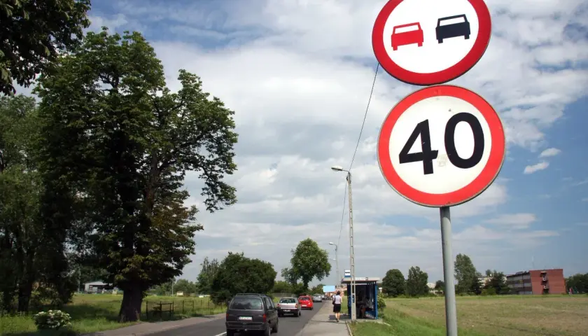 Znaki drogowe: zakaz wyprzedzania i ograniczenie prędkości do 40 km/h. Samochody jadą drogą, po prawej stronie przystanek autobusowy.
