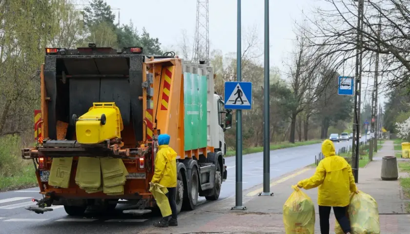 Śmieciarka i dwie osoby w żółtych płaszczach zbierające worki z odpadami.