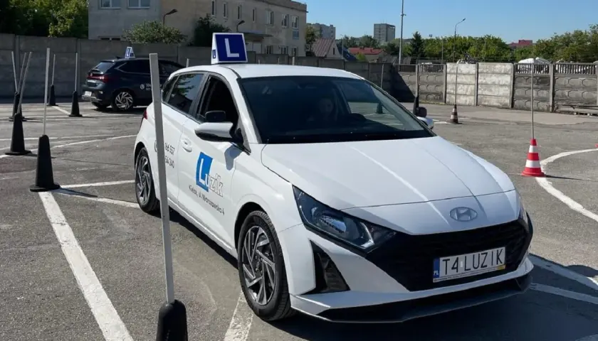Biały samochód nauki jazdy z literą "L" na dachu, na placu manewrowym.