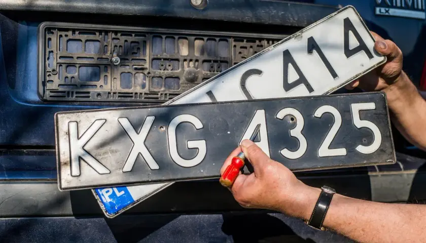 Wymiana tablic rejestracyjnych. Jedna tablica z numerem KXG 4325 jest przykręcana, druga z numerem C414 jest trzymana w ręku.