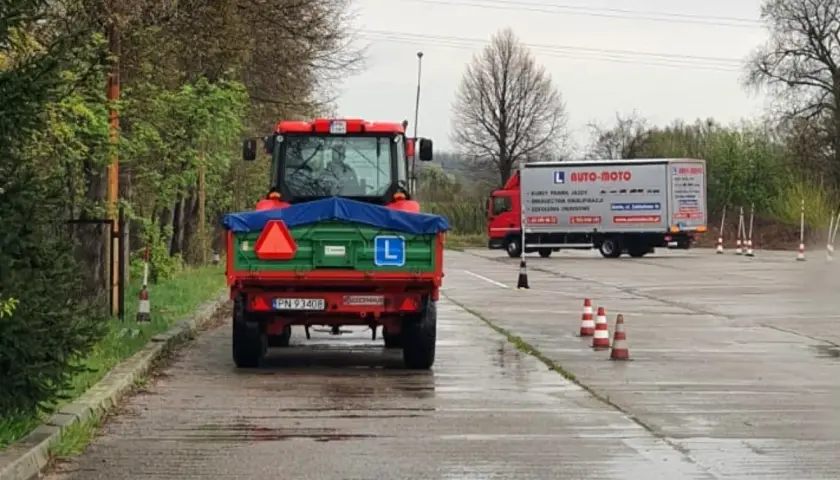 Czerwony traktor z przyczepą na placu manewrowym, obok ciężarówka AUTO-MOTO.