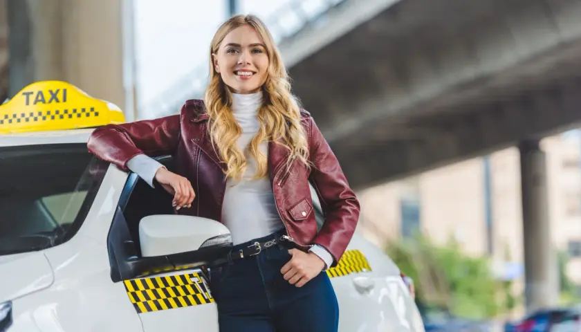Uśmiechnięta blondynka w skórzanej kurtce opiera się o białe taxi. Chcesz wiedzieć, jak zostać kierowcą Opti Taxi?