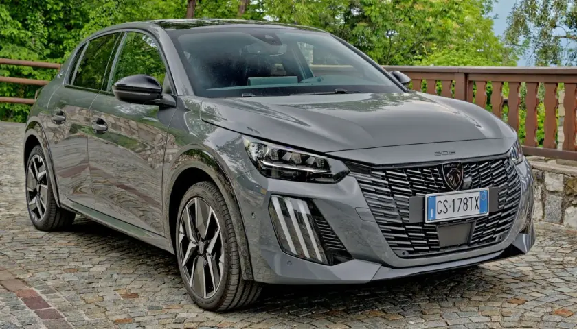 Szary Peugeot 208 zaparkowany na brukowanej drodze, z zielenią w tle.