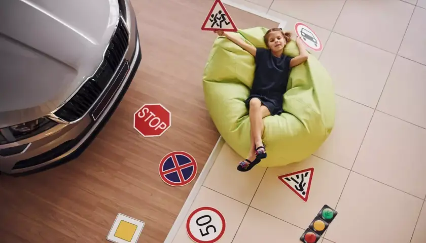 Dziewczynka z tabliczkami drogowymi, jakby uczyła się, ile jest godzin jazdy na prawo jazdy. Obok samochód i znaki: stop, dzieci, ograniczenie prędkości.