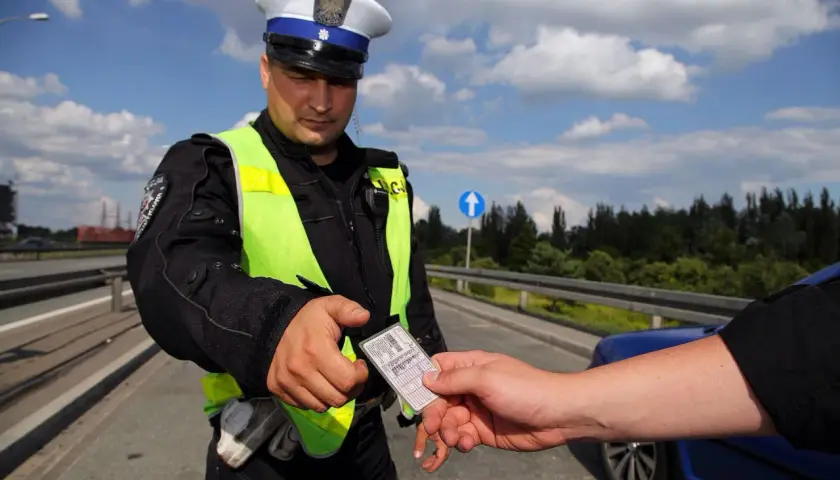 Policjant w kamizelce odblaskowej odbiera dokument. Czy trzeba mieć przy sobie prawo jazdy? Tak, podczas kontroli drogowej.
