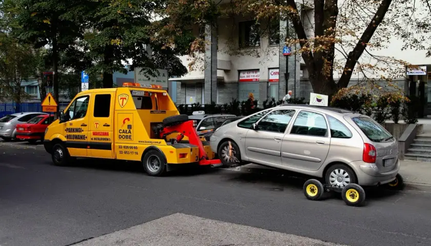 Żółta laweta holuje srebrny samochód, który zaparkował na zakazie. Mandat za parkowanie na zakazie jest nieunikniony.