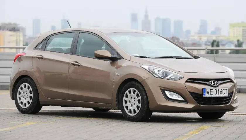 Brązowy Hyundai i30 1.4 benzyna na parkingu, w tle miejska panorama.