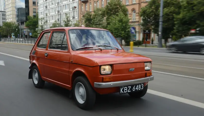 Pomarańczowy Fiat 126p jedzie ulicą. Jego pojemność baku to 25 litrów, co pozwala na długie podróże.