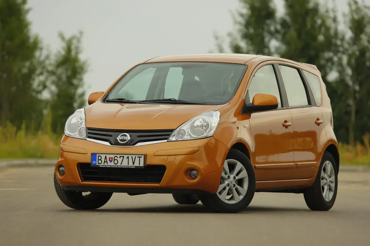 Pomarańczowy Nissan Note z przodu, na tle drzew.