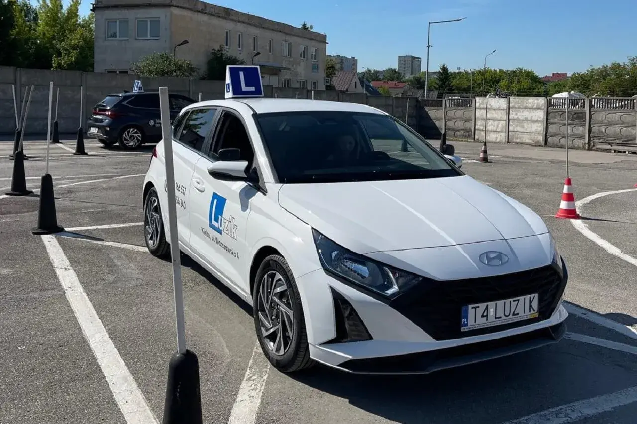 Biały samochód nauki jazdy z literą "L" na dachu, na placu manewrowym.