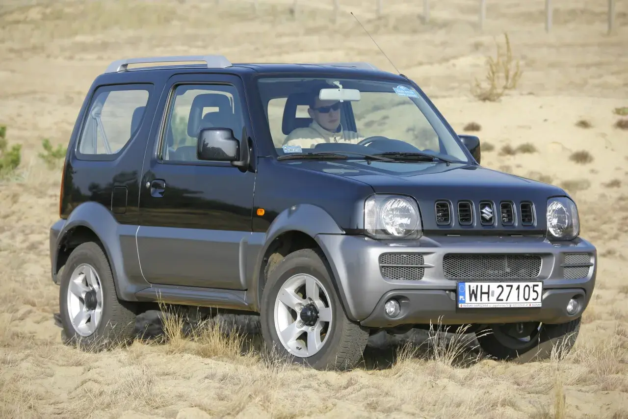 Czarny Suzuki Jimny na piasku. Kierowca w okularach. Pojemność baku tego modelu jest idealna na długie wyprawy.