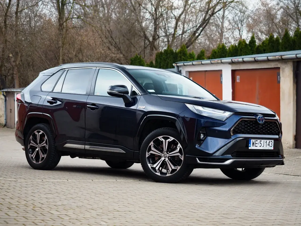 Ciemnoniebieski SUV Toyota RAV4 hybrydowy zaparkowany przed garażami.