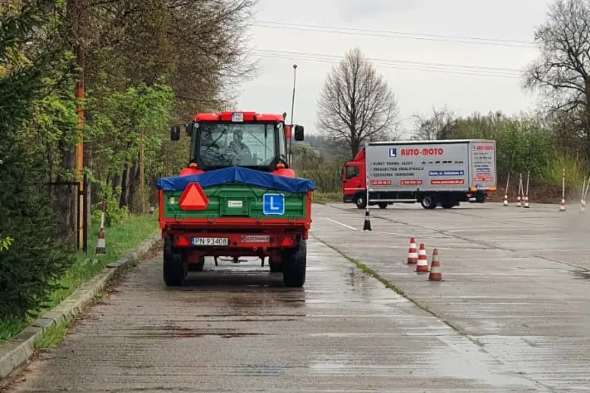 Czerwony traktor z przyczepą na placu manewrowym, obok ciężarówka AUTO-MOTO.