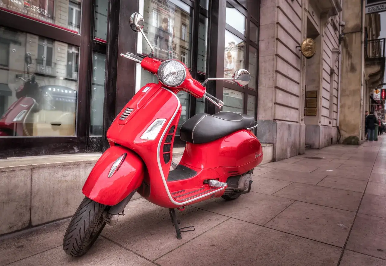 Czerwony skuter Vespa, idealny do zdobycia **prawo jazdy AM**, stoi na chodniku przed budynkiem.