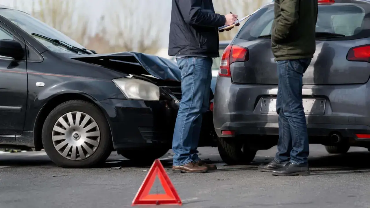 Dwaj mężczyźni spisują protokół po kolizji. Zniszczona maska czarnego auta i czerwony trójkąt ostrzegawczy sugerują, że czeka ich mandat za kolizje.